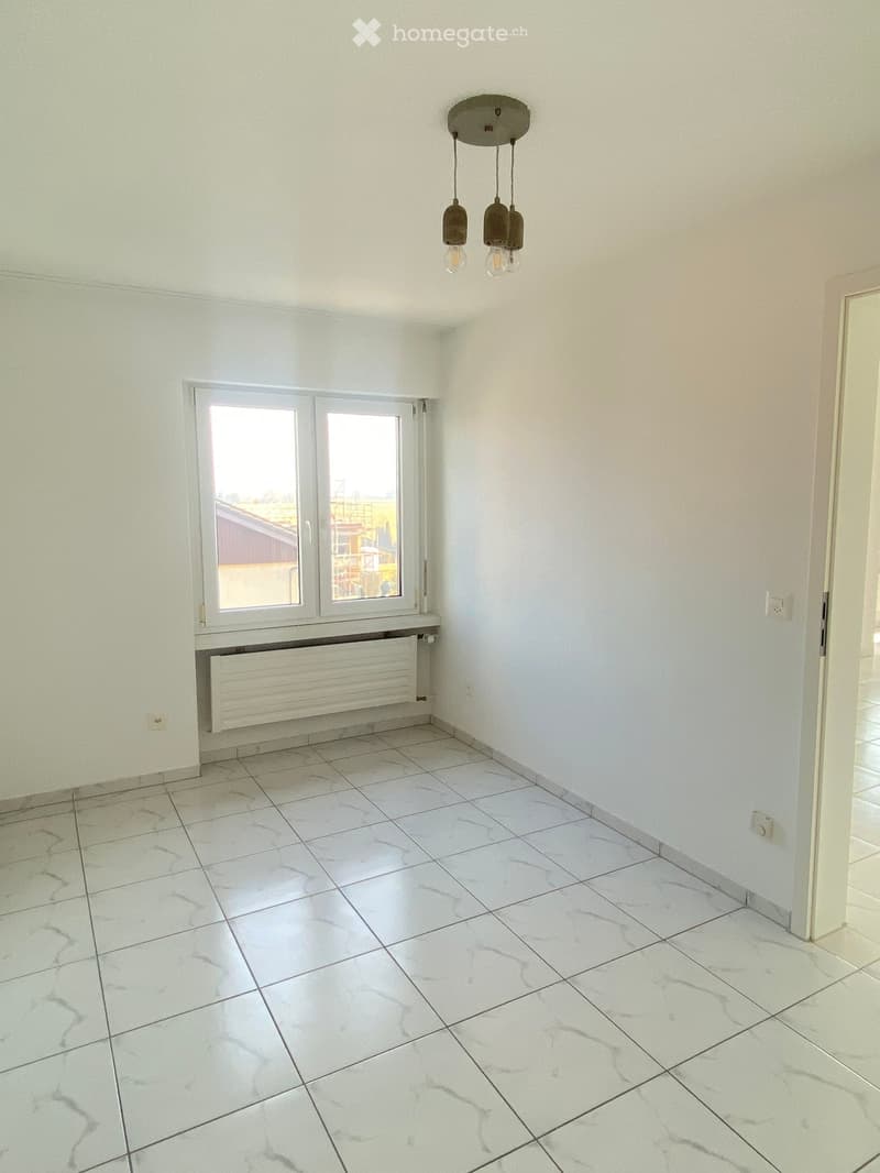 Empty room with white tiled floor, white walls, large window with view, radiator, and hanging light fixture.