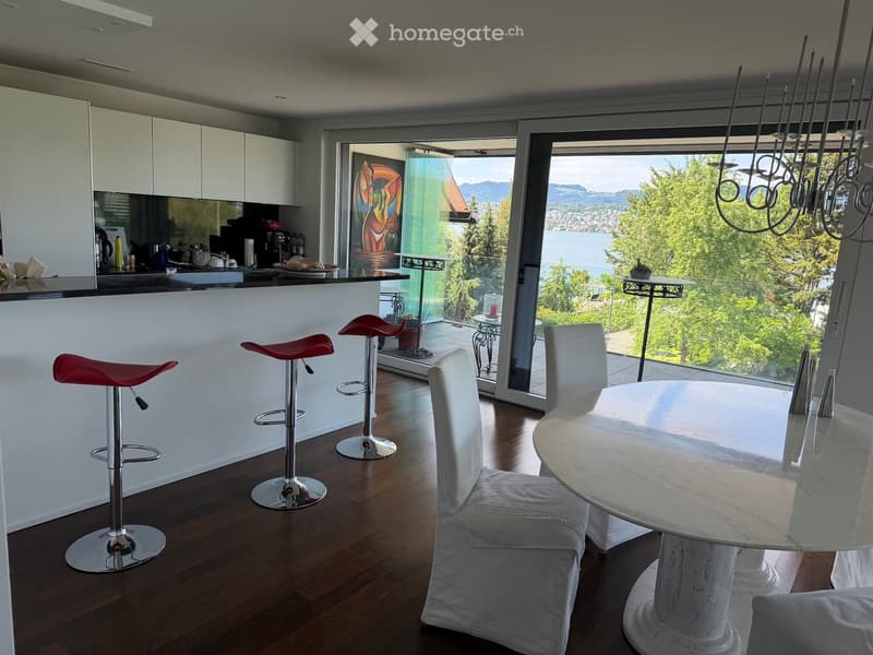 Modern kitchen and dining area with white cabinets, a black countertop, red bar stools, a white dining table with chairs, and a view of a lake and mountains through large windows.