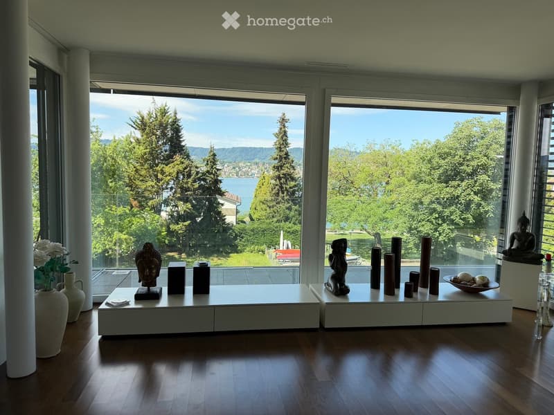 Bright living room with large windows offering a scenic view of trees and water. Decor includes white tables, vases, sculptures, and a bowl with decorative items.