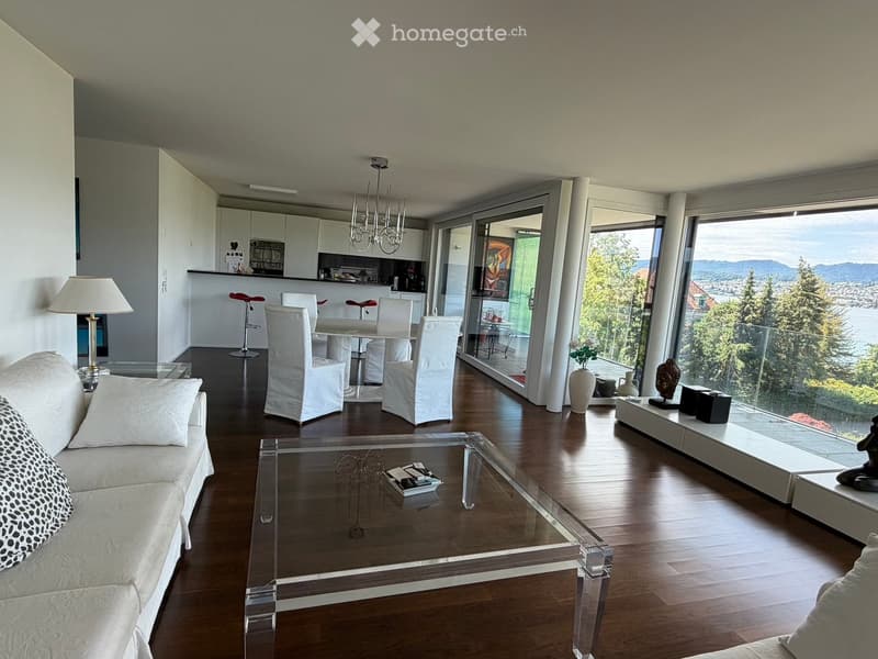 Modern living room with a sofa, coffee table, dining area with chairs and stools, large windows offering a scenic view, and decorative items including a vase and statue.