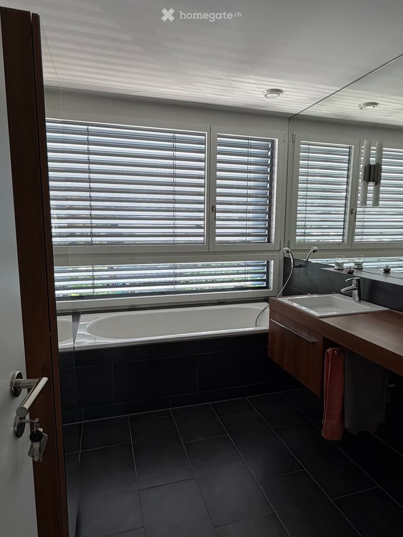 Modern bathroom with a large window, bathtub, double sink, and mirrored cabinet. Dark tiled floor and walls, sleek wooden vanity, and a towel hanging on the side.