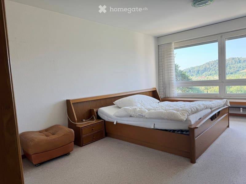 Bedroom with a wooden bed, nightstand, and a small brown stool, large windows with a scenic view.