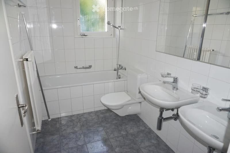 Modern bathroom with white tiled walls and grey tiled floor, featuring a bathtub, toilet, double sink, mirror, and radiator.