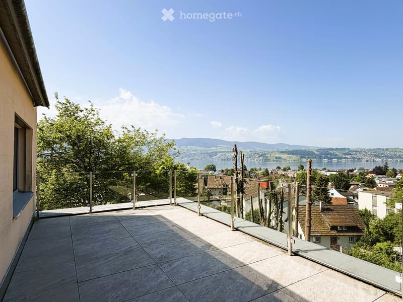 A modern balcony with a glass railing, overlooking a scenic town and lake with distant mountains.