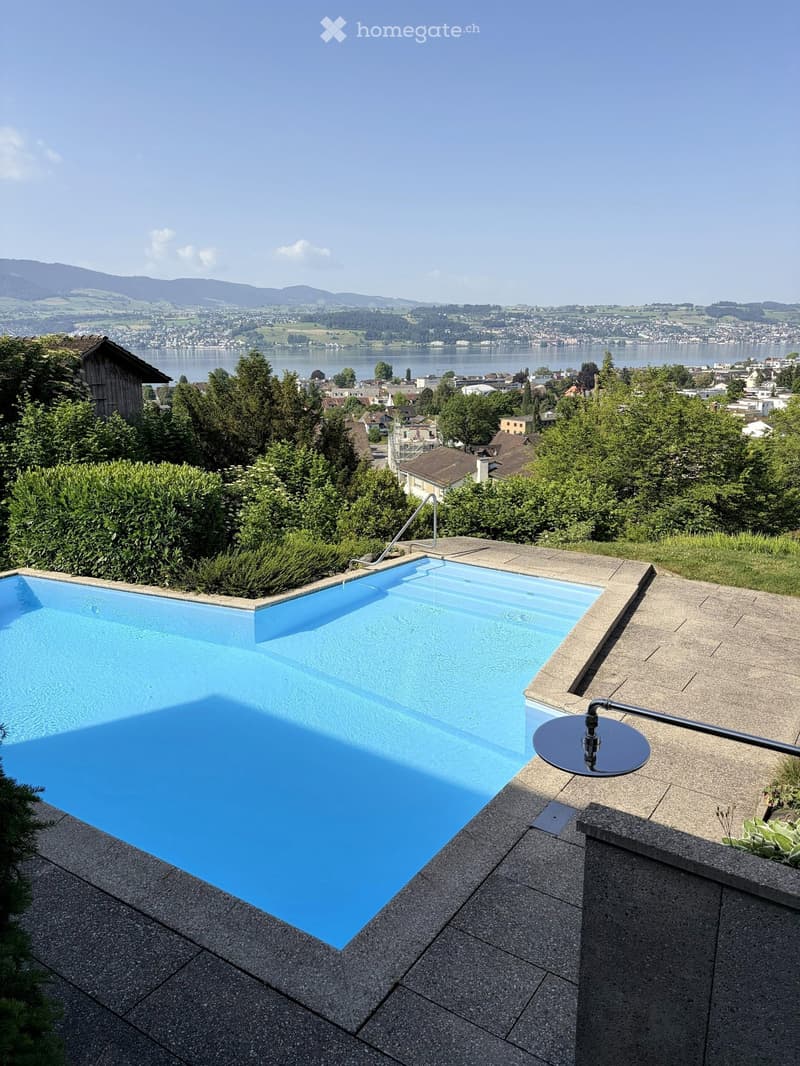 A modern swimming pool with a clear blue water surface, surrounded by a stone deck and lush greenery, overlooking a scenic lake and distant mountains.
