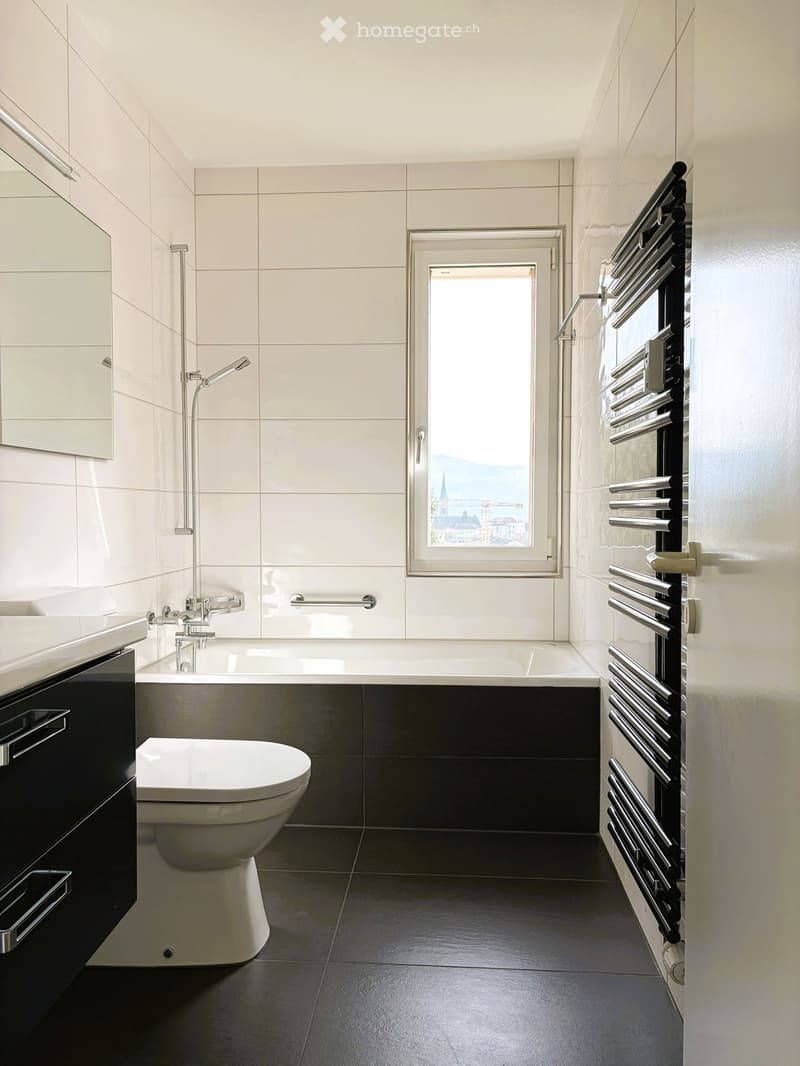 Modern bathroom with white tiled walls, dark floor tiles, a bathtub with a separate shower head, a toilet, vanity with sink, and a mirror mounted on the wall above the vanity.