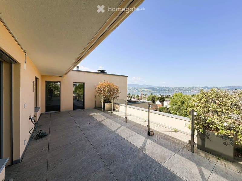 Spacious balcony with tiled flooring, modern glass railing, and scenic view of a lake and surrounding landscape.