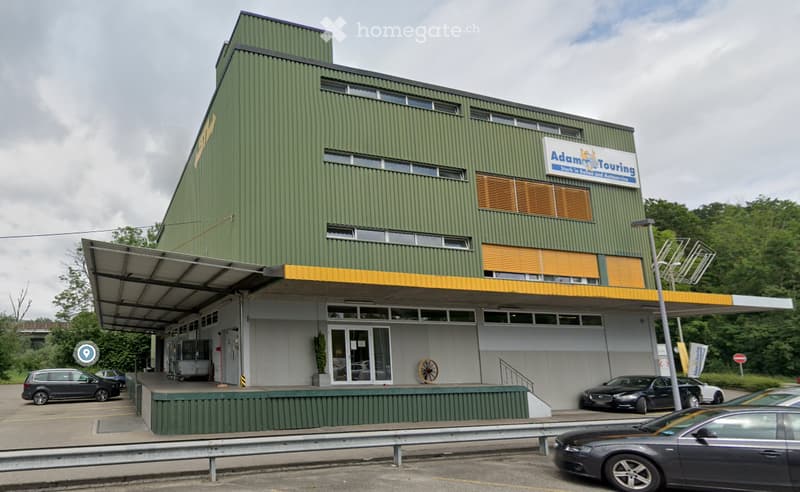 Modern green building with a sign reading "Adam Touring" and a covered entrance area. Several cars are parked in front.