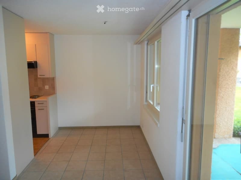Empty room with tiled floor, white walls, and a sliding glass door leading to a balcony. A small kitchen area is visible in the background.