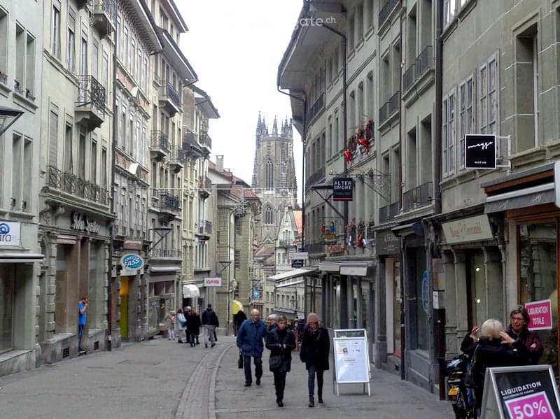 A bustling urban street lined with multi-story buildings, featuring shops, cafes, and pedestrians. The architecture includes ornate facades and balconies, with a cathedral visible in the background.