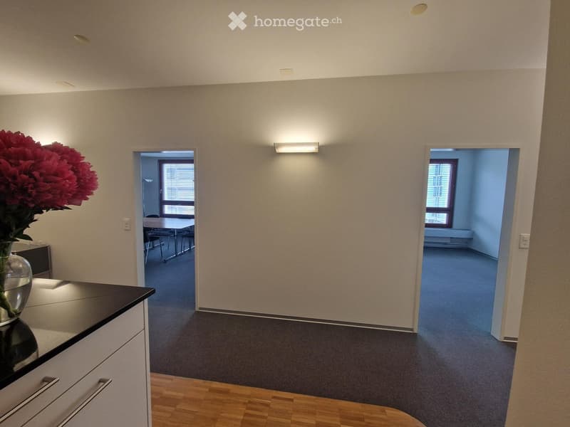 A well-lit hallway with beige walls, wooden flooring, and two open doorways leading to adjacent rooms. One room contains a table and chairs, while the other is empty with blue walls and carpeted flooring.