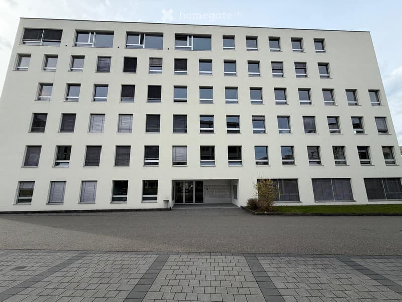 Modern, multi-story white building with numerous windows and a symmetrical facade, featuring a central entrance and paved ground area.