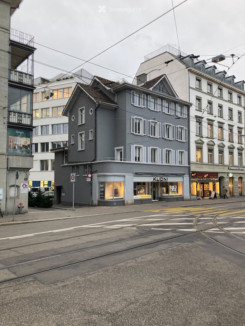 A multi-story residential building with grey facade, multiple windows, and commercial storefronts including 'KUONI' and 'STARBUCKS COFFEE'.