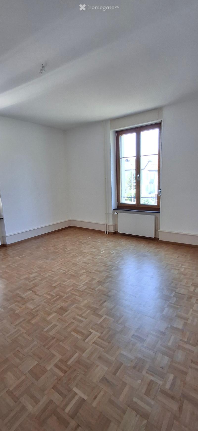 Empty room with wooden herringbone flooring, white walls, and a large window with brown frame. A radiator is visible beneath the window.