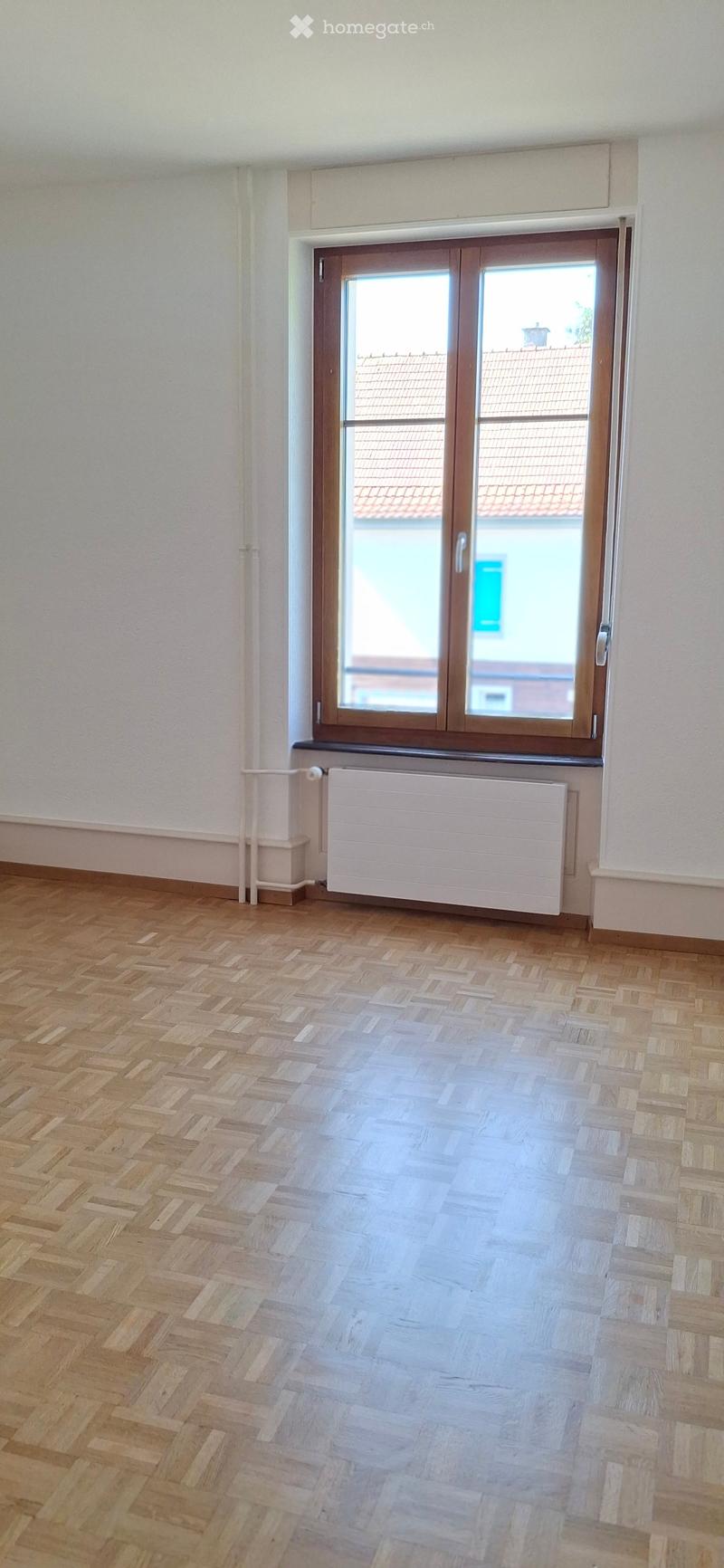 Empty room with wooden herringbone flooring, white walls, a radiator, and a large window with a view of a neighboring building.