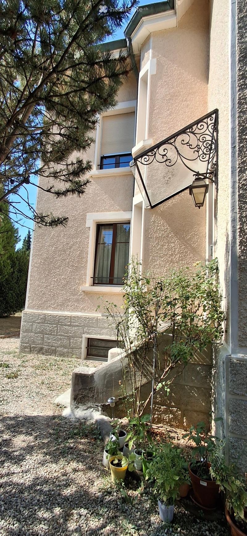 Two-story house with beige stucco exterior, multiple windows, decorative wrought-iron balcony, and small garden area with potted plants and gravel ground.