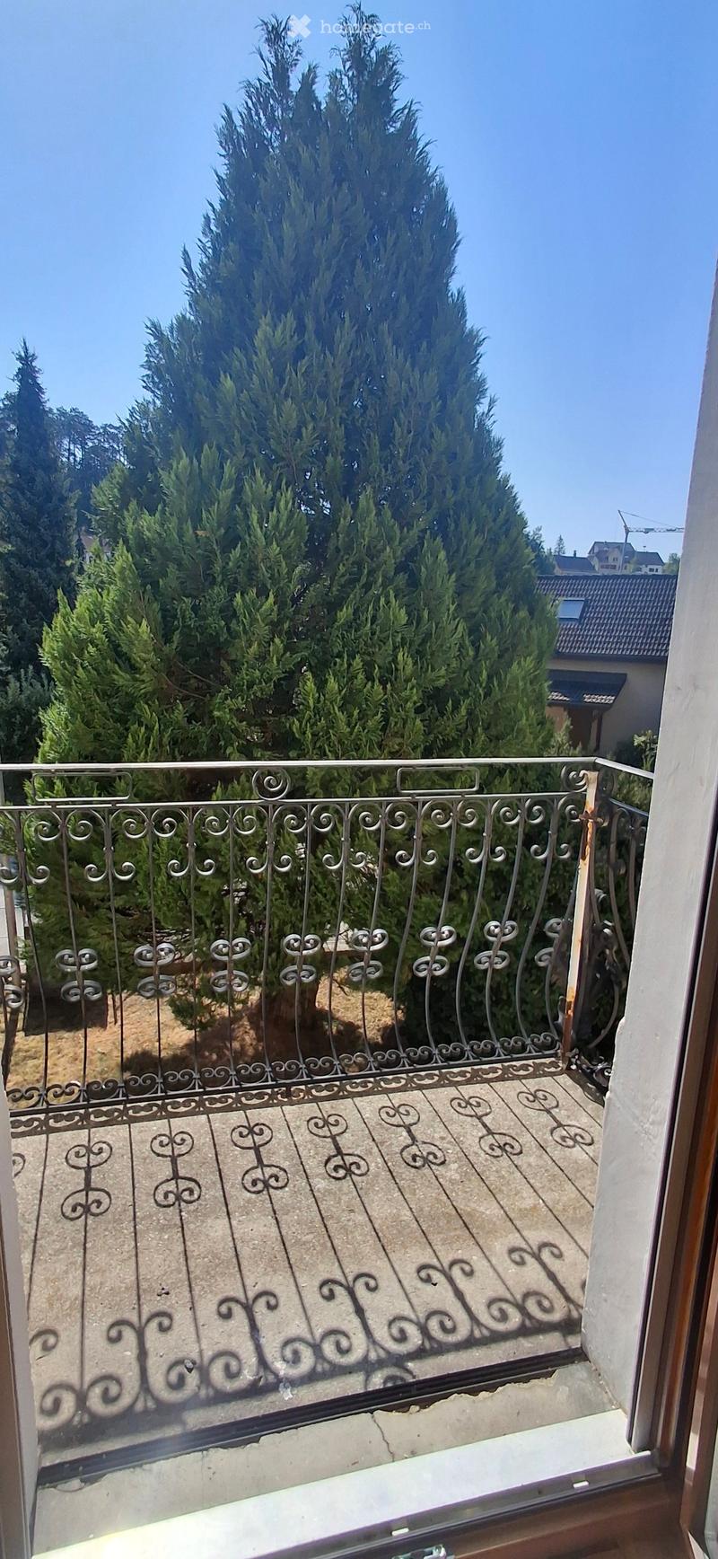 Balcony with ornate metal railing, tiled floor, and view of trees and neighboring houses.