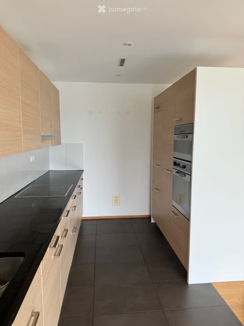 Modern kitchen with light wood cabinets, black countertop, and built-in oven. The space features a clean, minimalist design with white walls and dark tiled flooring.