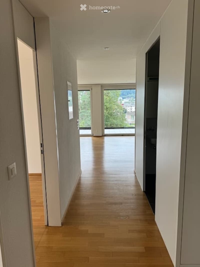 Bright hallway with wooden flooring, white walls, large windows, and a view of greenery outside.