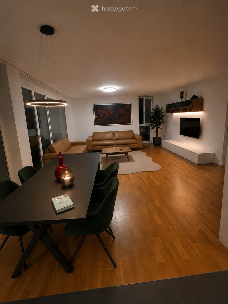 Modern living and dining area with wooden flooring, a large sofa, coffee table, dining table with chairs, decorative vases, a candle, and a TV mounted on the wall.