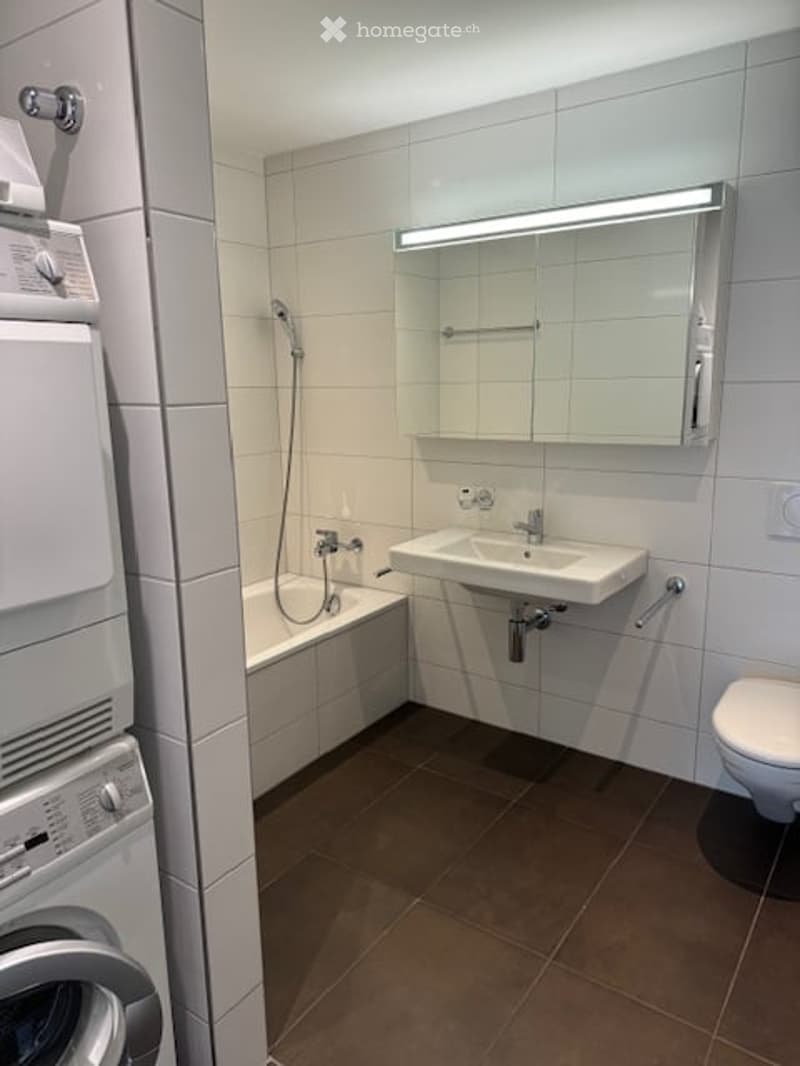 Modern bathroom with white tiles, a bathtub, a sink with a mirror, a toilet, and a washing machine.