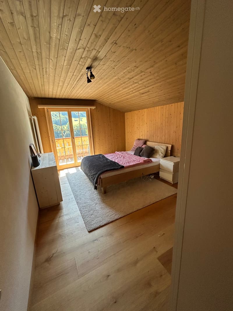 Cozy attic bedroom with wooden walls and ceiling, large window with view, bed with pillows and blankets, nightstand, and lamp.