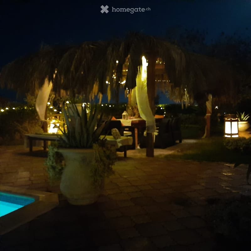 Nighttime outdoor area with a swimming pool, stone-paved ground, large thatched-roof structure, tables and chairs, decorative lanterns, and potted plants.