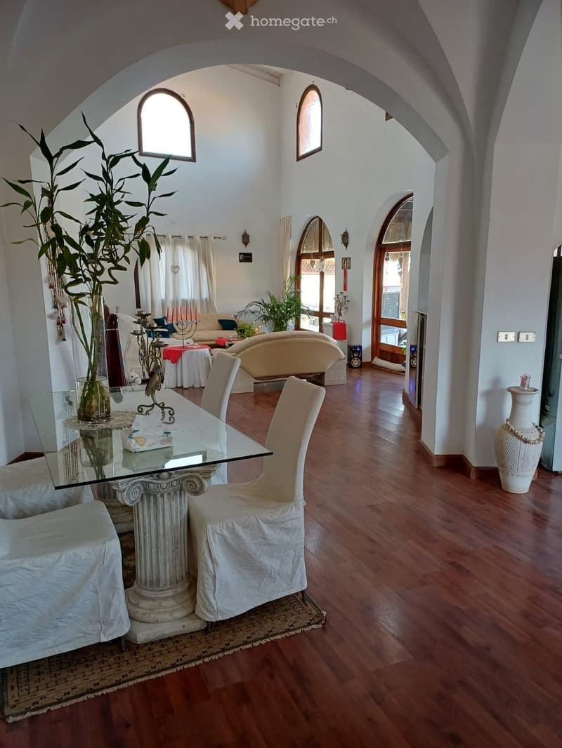 Elegant living and dining area with wooden flooring, white walls, arched doorways, and large windows. Features a glass dining table with four chairs, a cream-colored sofa, a decorative vase, and a tall plant in a pot.