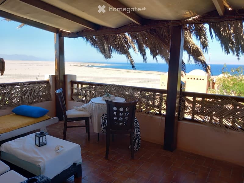 Balcony with a thatched roof, overlooking the ocean, featuring a small table, chairs, a bed with pillows, and a lantern.