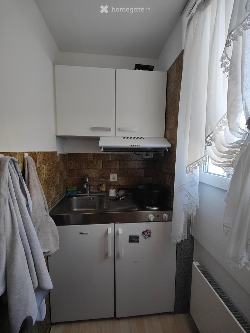 Compact kitchen with white cabinets, stainless steel sink, refrigerator, and small oven. Wooden floor and tiled backsplash.