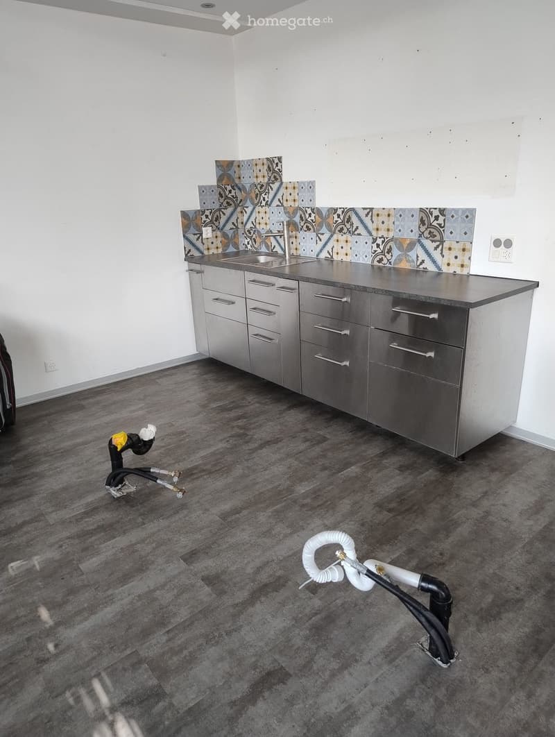 Empty kitchen with stainless steel cabinets, a sink, and a decorative tile backsplash. The floor is made of dark wood planks.