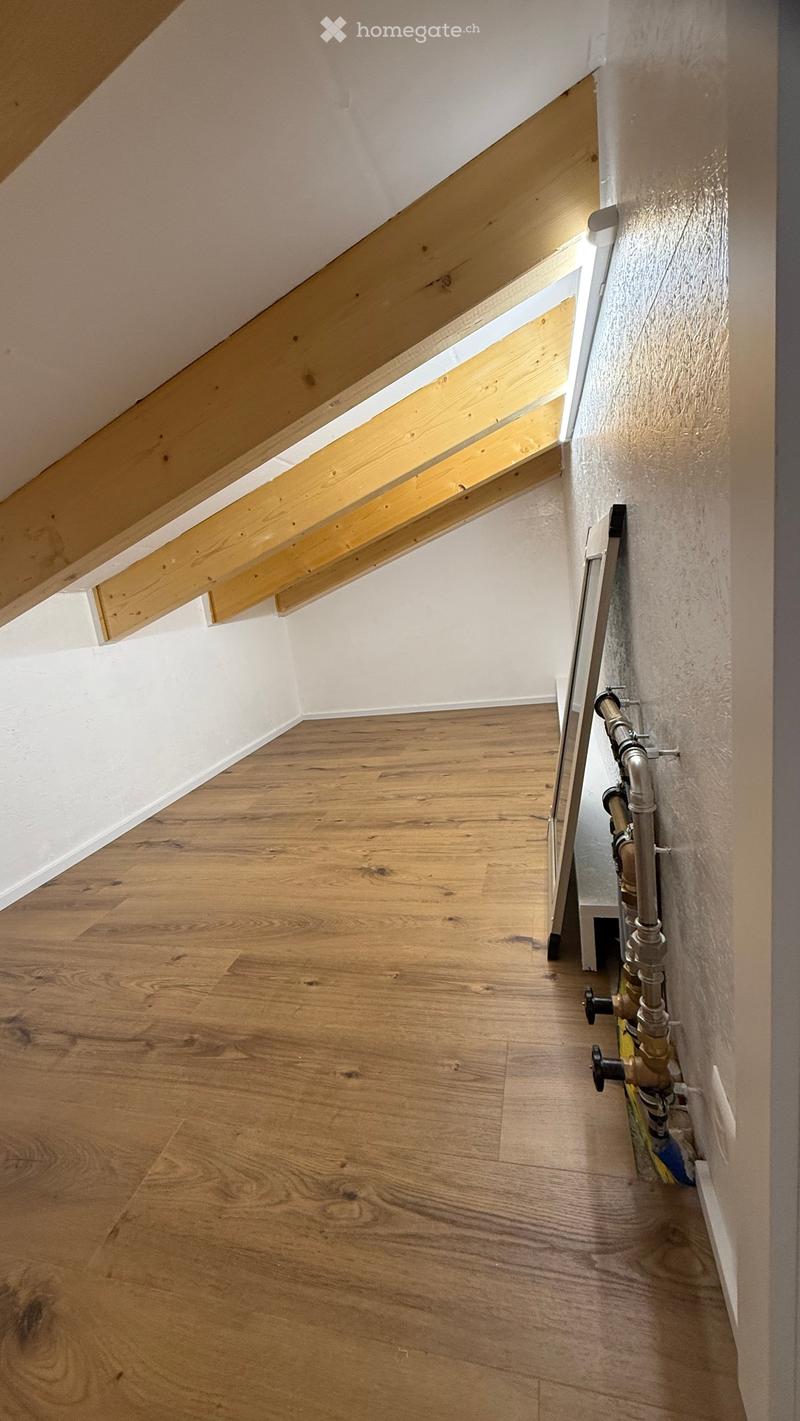 Empty room with wooden flooring, white walls, and exposed wooden beams in the ceiling. Plumbing fixtures are visible on one wall.