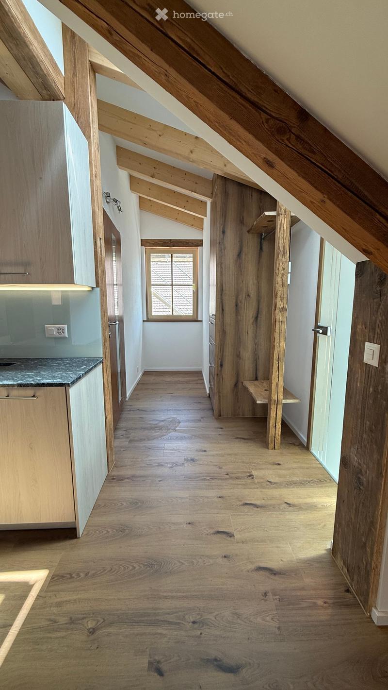 Bright, wooden-floored hallway with exposed wooden beams and a small window at the end, leading to a kitchen area with light wood cabinets and a granite countertop.