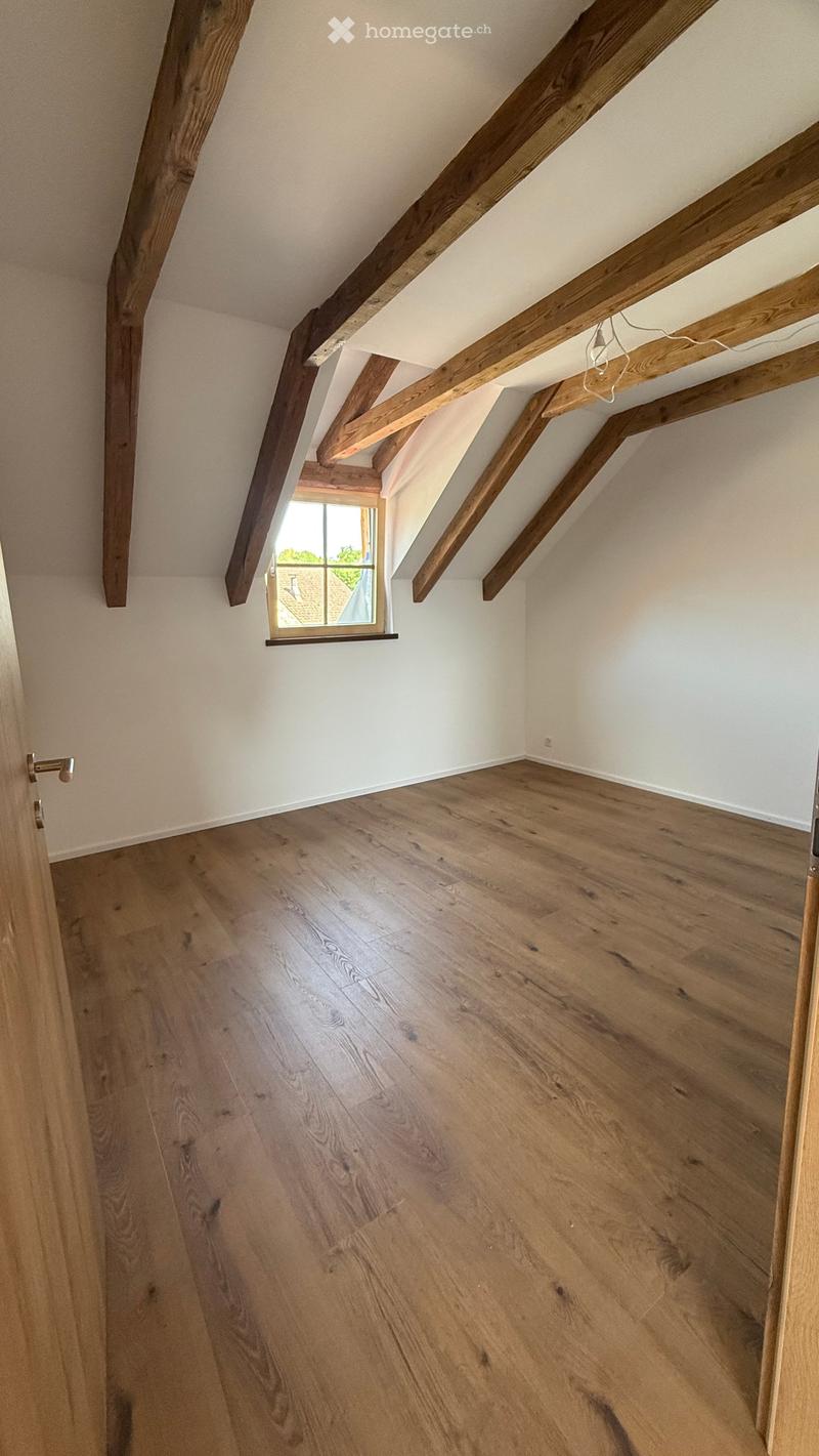 Empty attic space with exposed wooden beams, wooden flooring, and a small window letting in natural light.