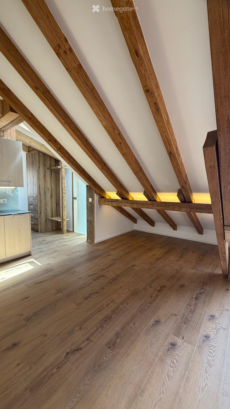 An attic space with exposed wooden beams and a wooden floor, featuring a small built-in shelf and a doorway leading to another room.