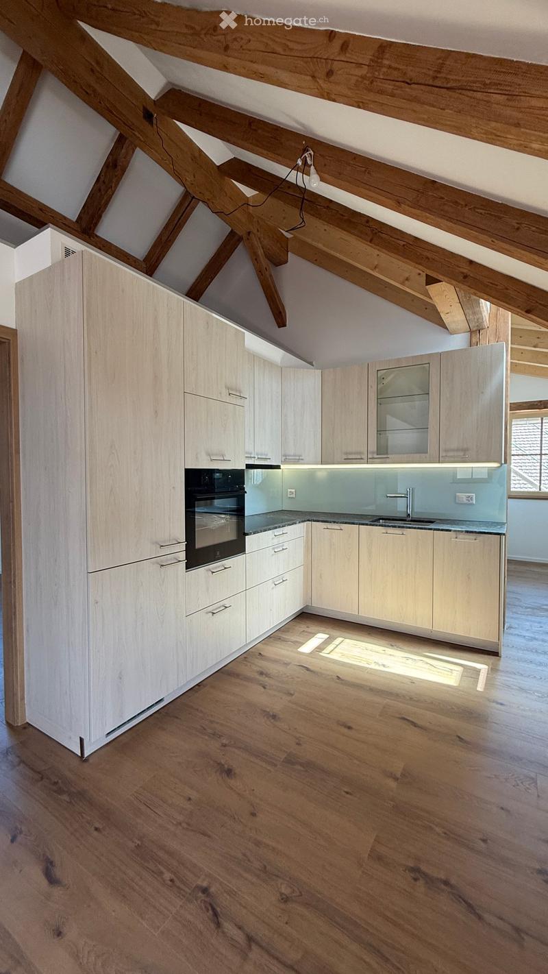 Modern kitchen with light wood cabinets, stainless steel oven, and a sleek countertop, featuring exposed wooden beams on the ceiling and a polished wooden floor.