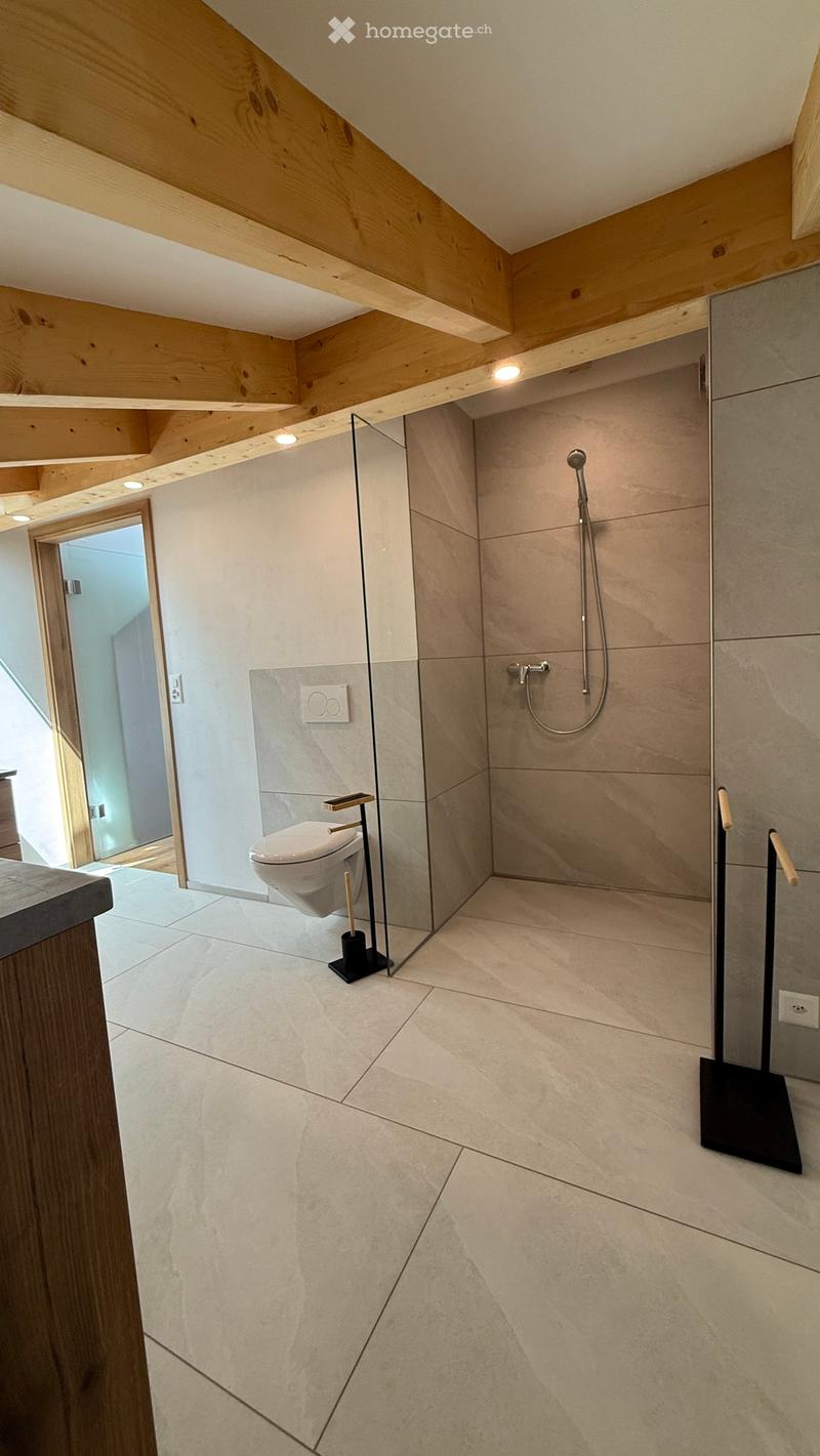 Modern bathroom with light-colored tiles, a glass-enclosed shower area, a toilet, and a sleek shower hose. The ceiling features exposed wooden beams and recessed lighting.