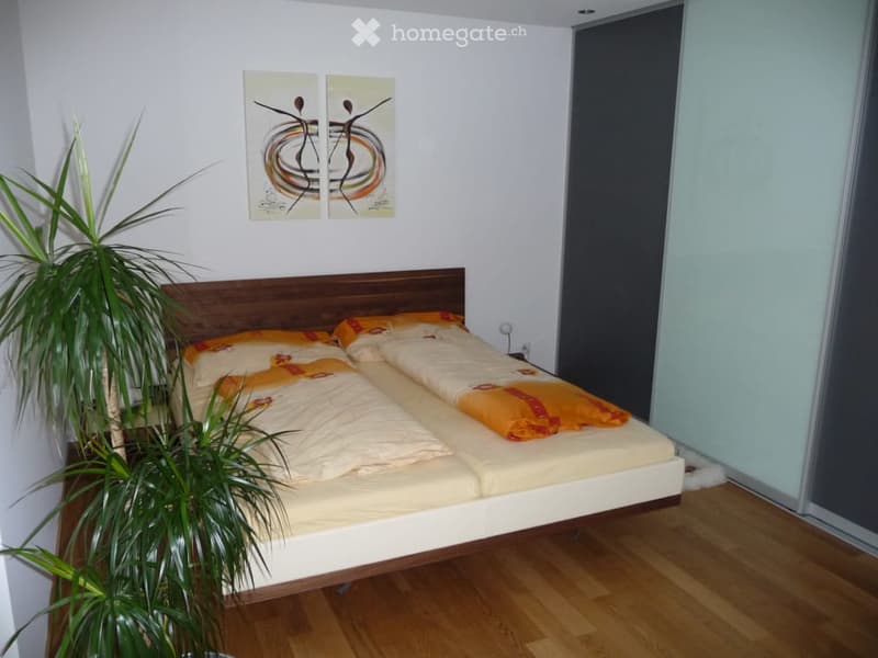 Bright bedroom with a double bed, wooden headboard, and two pillows, complemented by a nightstand and a potted plant near the window.