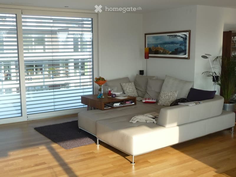 Modern living room with large windows offering a city view, featuring a white sectional sofa, wooden coffee table, floor lamp, picture frame, and a potted plant.