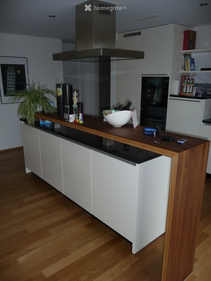 Modern kitchen with a wooden island, white cabinets, and a stainless steel range hood. The island features a dark countertop with a bowl, plant, and various small items.
