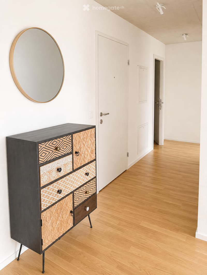 Modern hallway with light wooden flooring, white walls, a large round mirror with a gold frame, and a dark wooden cabinet with patterned drawers.