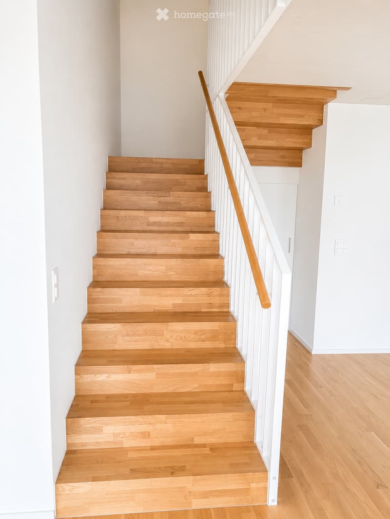 Wooden staircase with white railings and a wooden handrail, leading up to a landing area with light-colored walls and wooden flooring.