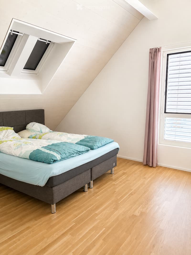 A cozy attic bedroom with wooden flooring, a large bed with a grey headboard, colorful bedding, and a window with blinds and curtains. The room features skylight windows in the sloped ceiling.
