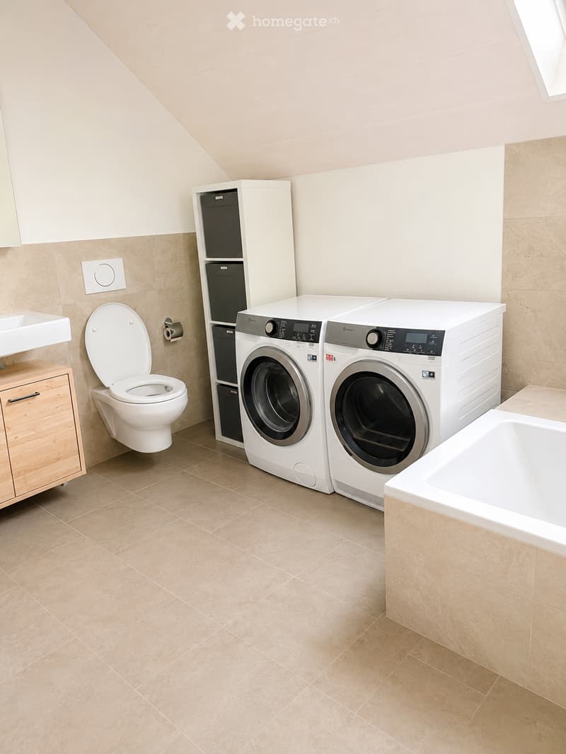 Modern bathroom with beige tiles, a toilet, sink, bathtub, washing machine, and dishwasher.