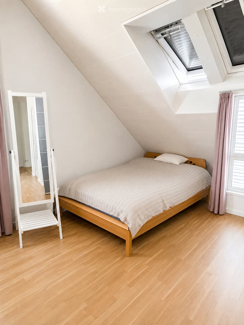 Cozy attic bedroom with wooden flooring, slanted ceilings, and skylight windows. Features a wooden bed with striped bedding, a white mirror, and a small white stool.