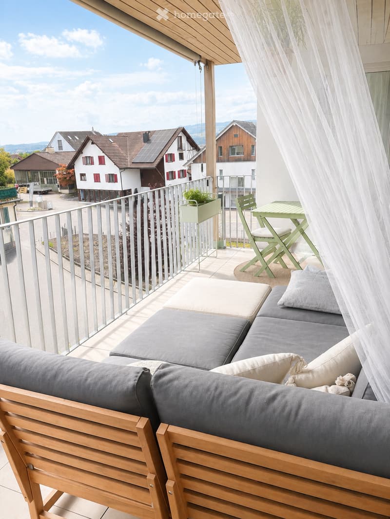 Balcony with wooden sofa, grey cushions, small table, and chair, overlooking a residential area with houses and mountains.