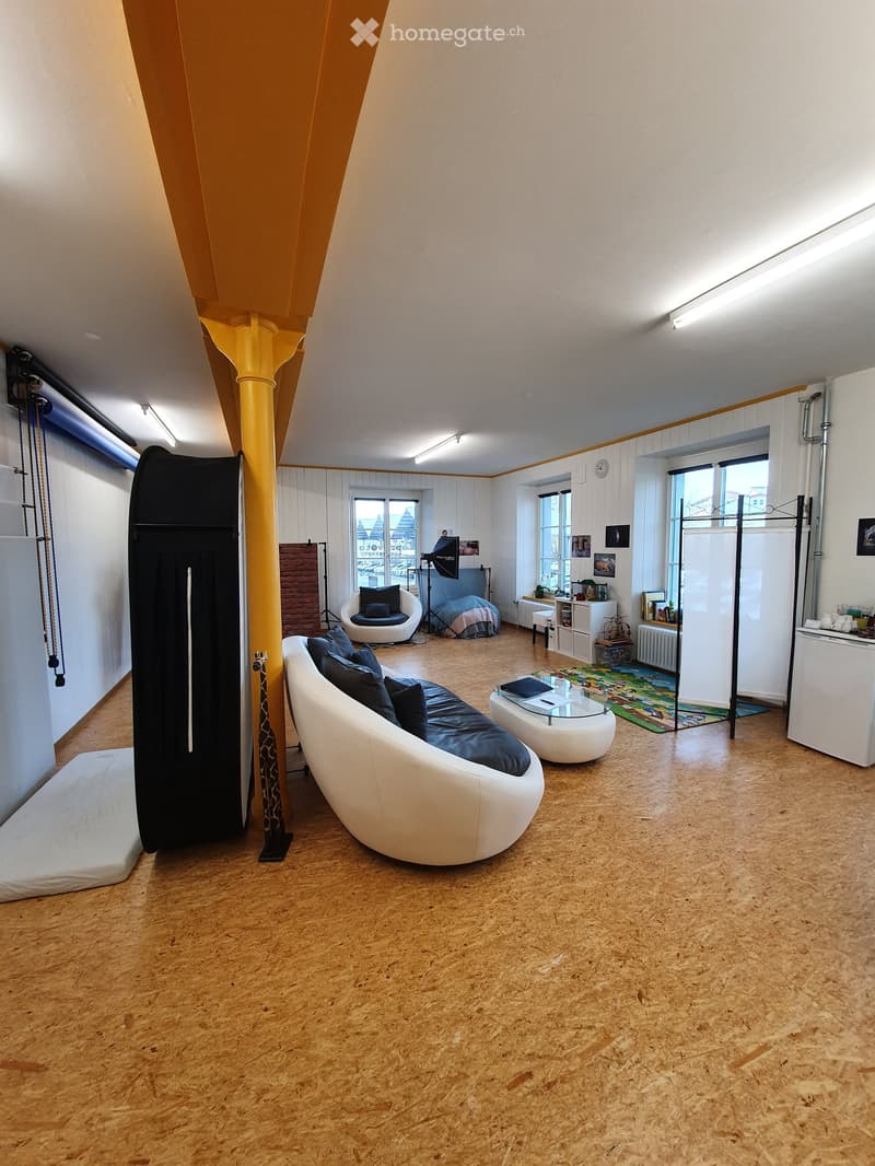 Modern living room with wooden flooring, white walls, large windows, a white sofa with blue cushions, a glass coffee table, a white chair, a screen, and a small table with a radiator.