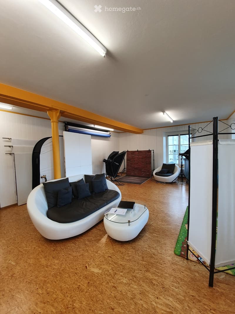 Modern living room with a white, curved sofa, glass coffee table, and a white egg chair. The room features wooden flooring, exposed beams, and a large window with a view.