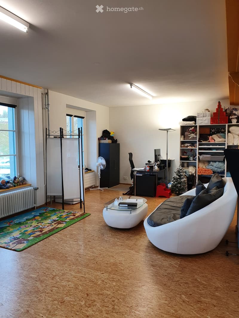 A modern living room with a white leather sofa, glass coffee table, desk, and shelving unit. The room features wooden flooring, large windows, and a colorful children's play mat.