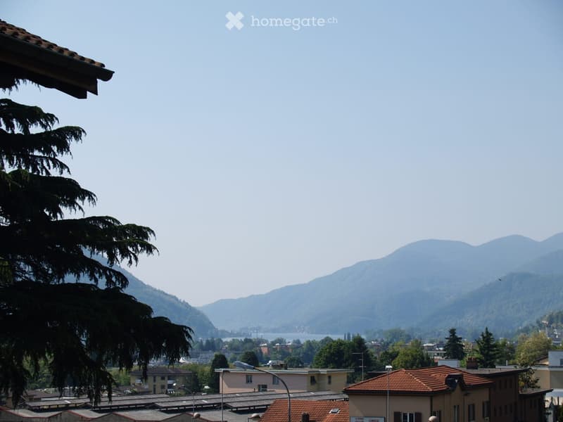 View from a balcony showing a cityscape with buildings, trees, and distant mountains under a clear blue sky.